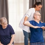 A physiotherapist assisting an elderly man with arm exercises while an elderly woman performs seated exercises in a home setting, featuring comfortable surroundings and fitness equipment in the background.