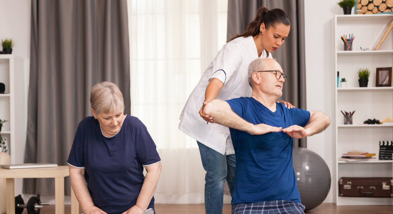 A physiotherapist assisting an elderly man with arm exercises while an elderly woman performs seated exercises in a home setting, featuring comfortable surroundings and fitness equipment in the background.