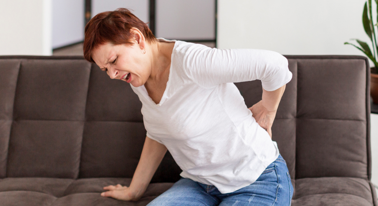Woman sitting on a couch, grimacing in pain while holding her lower back.