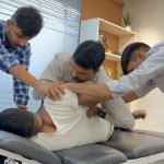 A team of three physiotherapists working together to perform advanced manual therapy on a patient lying on a treatment table in a modern clinic. The therapists are focused on adjusting the patient's upper body and hip area to improve mobility and relieve pain.