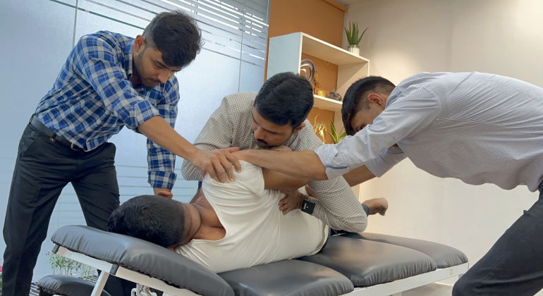 A team of three physiotherapists working together to perform advanced manual therapy on a patient lying on a treatment table in a modern clinic. The therapists are focused on adjusting the patient's upper body and hip area to improve mobility and relieve pain.