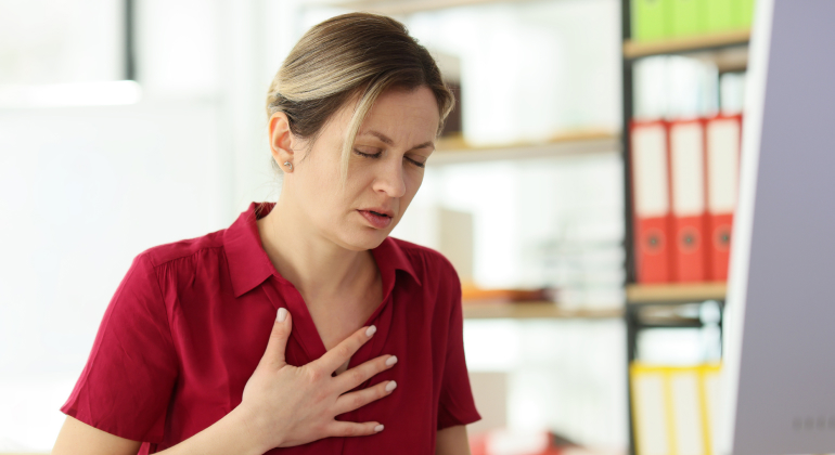 Woman in a red shirt clutching her chest in discomfort while standing in an office setting.