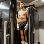 A man is standing inside a cryotherapy chamber. He is shirtless, wearing black shorts, white gloves, and socks, with his eyes closed, appearing calm and relaxed as the treatment begins. The chamber is sleek and modern, with a digital screen on the side displaying settings and controls. The room has a minimalist design with neutral tones, enhancing the clinical yet comfortable atmosphere.
