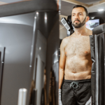 The image shows a man undergoing a whole-body cryotherapy session. He is standing inside a cryotherapy chamber, with the top half of his body exposed while the lower half is enclosed within the chamber. The man is wearing black shorts and appears calm and focused as he experiences the extreme cold temperatures used in cryotherapy. The chamber is sleek and modern, equipped with control panels and displays, highlighting the advanced technology used in cryotherapy treatments. This image captures the process of cryotherapy, emphasizing its role in wellness and recovery therapies.