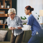 Physiotherapist assisting an elderly woman with a walking stick during a home physiotherapy session in a comfortable living room setting.