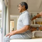 Physiotherapist performing back therapy on an elderly man during a home physiotherapy session in a bright, comfortable living space.