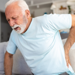 An elderly man sitting on a sofa holding his lower back in pain. He appears to be experiencing discomfort, indicating possible back or joint issues.