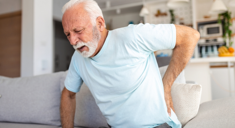 An elderly man sitting on a sofa holding his lower back in pain. He appears to be experiencing discomfort, indicating possible back or joint issues.