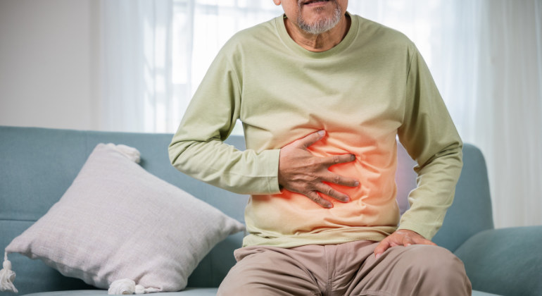 Elderly man sitting on a couch, holding his stomach in pain, with a red glow effect on the abdominal area, indicating digestive discomfort or stomach issues.
