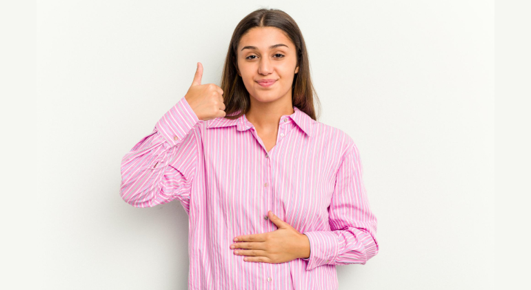 Young woman in a pink shirt smiling with one hand on her stomach and the other showing a thumbs up, symbolizing improved digestion from navel seed oil