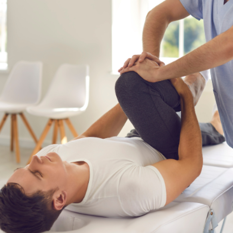 A physiotherapist performing a knee therapy session on a male patient lying on a treatment table in a clinic setting. The therapist is gently bending the patient’s leg to improve mobility.