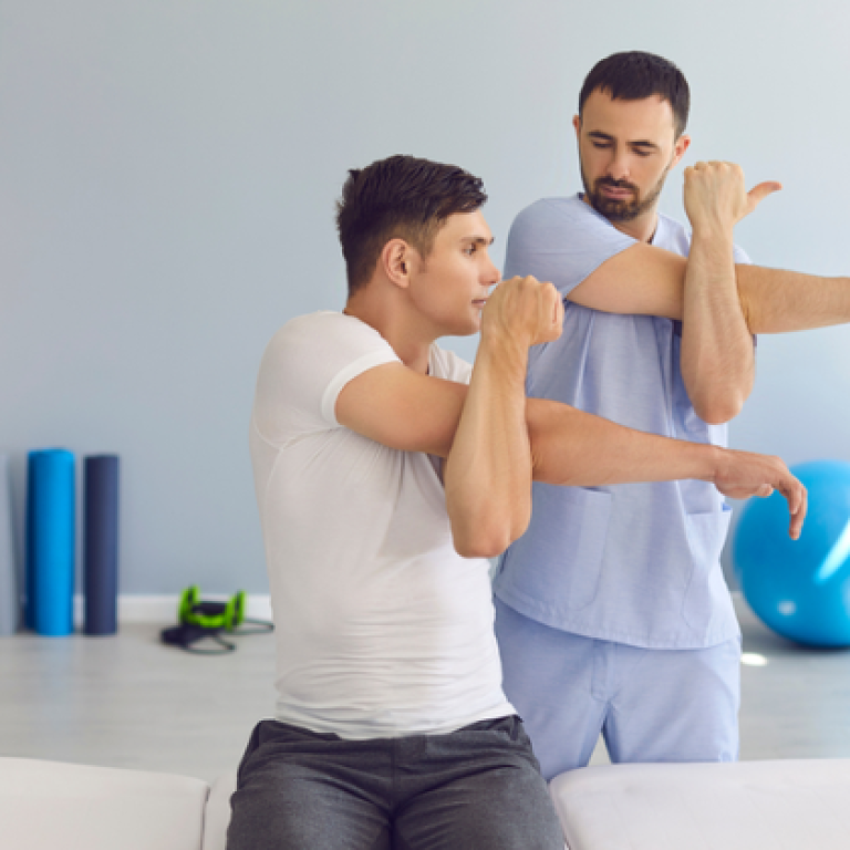 A physiotherapist guiding a male patient through shoulder stretching exercises. The patient is sitting on a treatment table, extending his arm across his chest in a clinic setting.