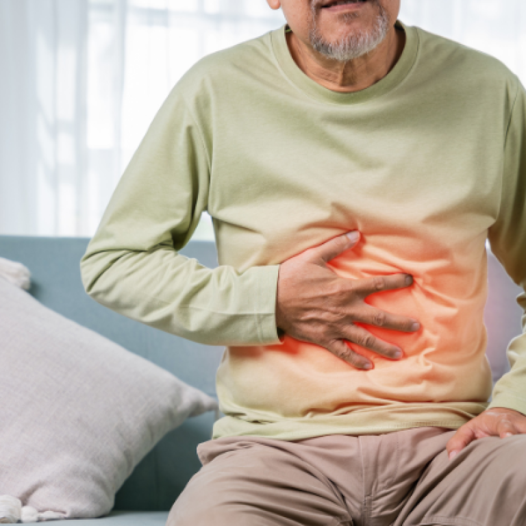 Elderly man sitting on a couch, holding his stomach in pain, with a red glow effect on the abdominal area, indicating digestive discomfort or stomach issues.