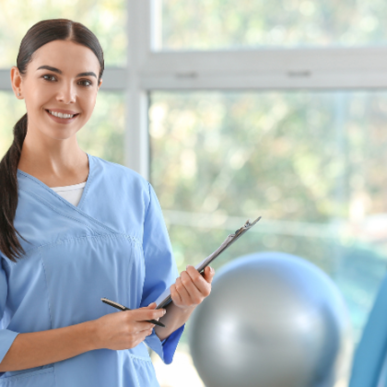 Smiling female physiotherapist in a light blue uniform holding a clipboard, standing in a bright, well-equipped therapy room with exercise equipment in the background.