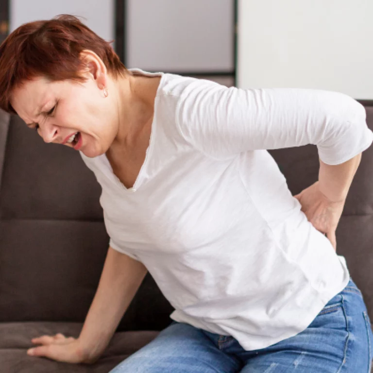 Woman sitting on a couch, grimacing in pain while holding her lower back.