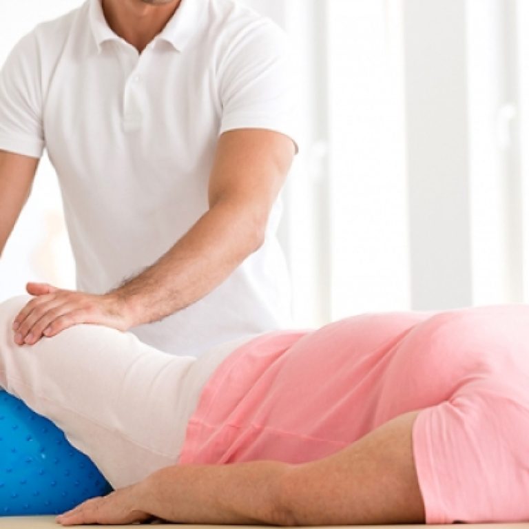 A physiotherapist assisting an elderly woman with a leg exercise during a home physiotherapy session. The woman is lying on her back with her legs bent over a large blue therapy ball. The therapist is supporting and guiding her leg movements to ensure proper technique and maximize benefits. The setting is a bright and comfortable home environment.