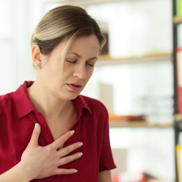 Woman in a red shirt clutching her chest in discomfort while standing in an office setting.