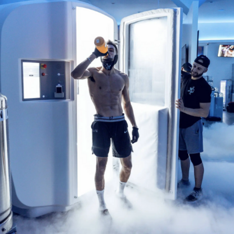 An athlete steps out of a cryotherapy chamber surrounded by cold vapor, wearing protective gloves and drinking from a bottle post-session. A technician assists nearby, with nitrogen tanks visible in the background used to cool the chamber for whole-body cryotherapy.