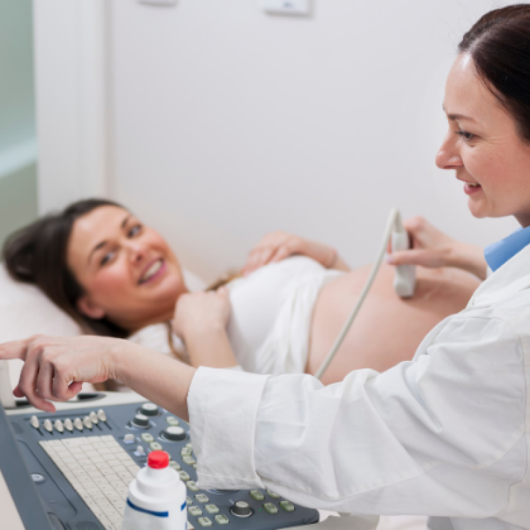 Pregnant woman lying on an examination bed smiling during an ultrasound scan, while a female doctor operates the ultrasound machine and explains the results.