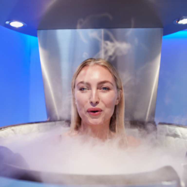 Person undergoing a cryotherapy session inside a chamber, with cold mist surrounding them in a blue-lit room.