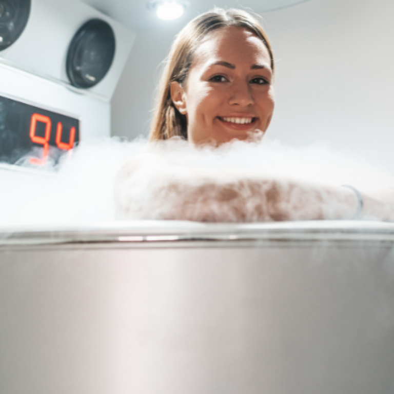 Smiling woman undergoing whole-body cryotherapy session in a cryotherapy chamber surrounded by cold mist.
